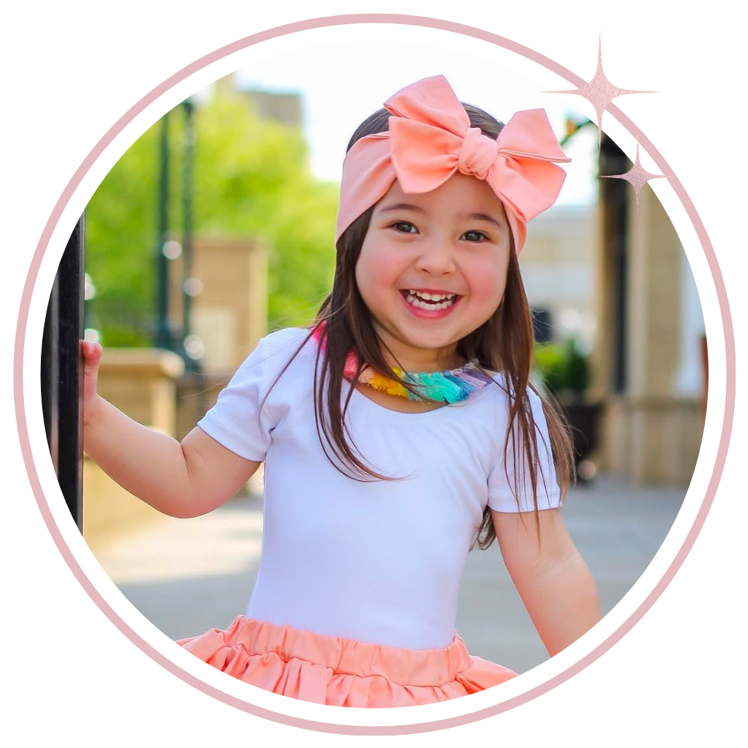 A young girl smiling and wearing a white short-sleeve top, a coral pink ruffled skirt, a matching coral pink bow headband, and a colorful feather necklace. The photo is framed in a circular border with sparkles on a white background.